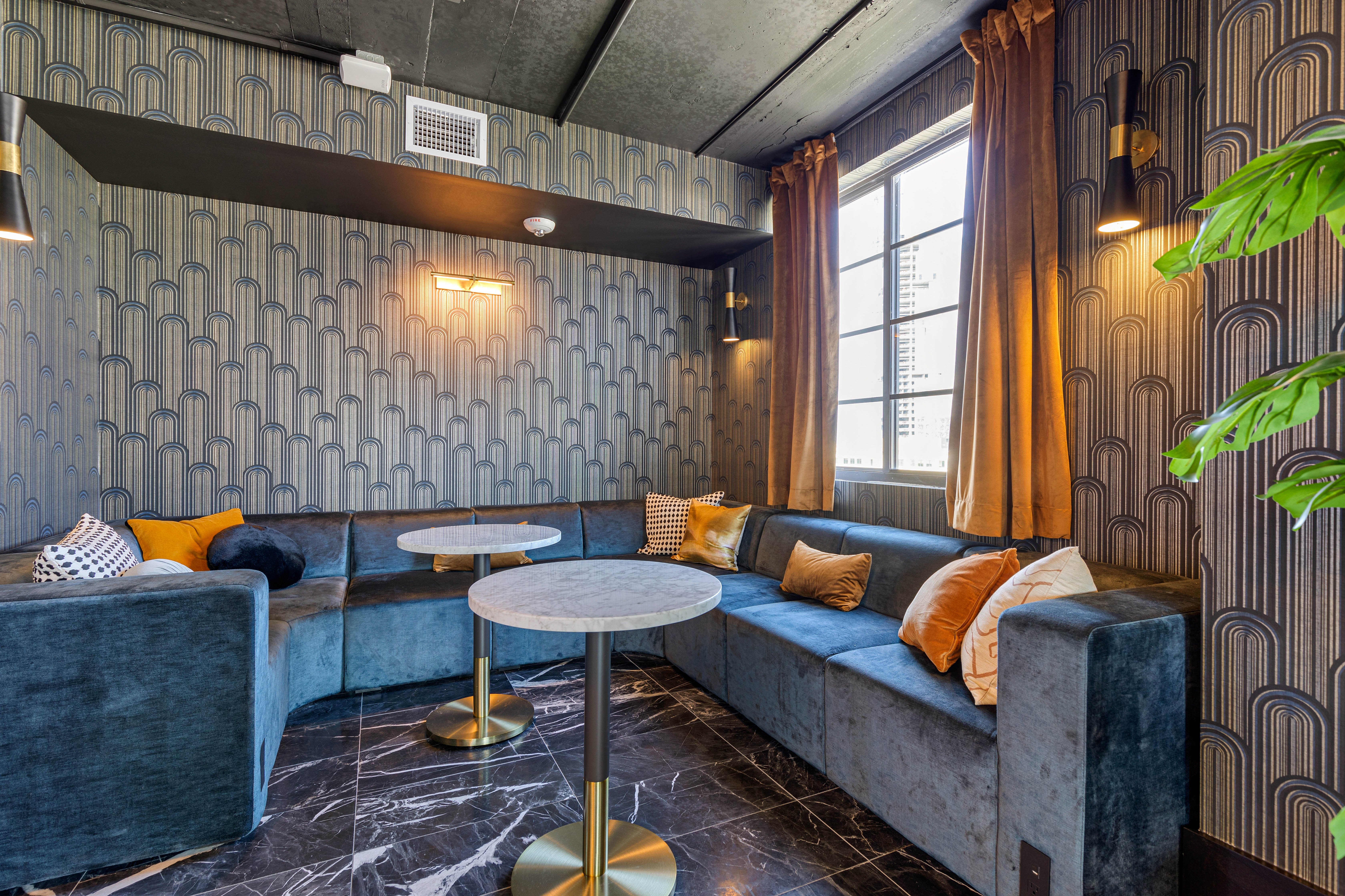 Contemporary Lounge Design A modern lounge area featuring a blue velvet sectional sofa with decorative cushions, two round marble tables, and large windows dressed with yellow curtains. The walls are adorned with a geometric pattern, and there are elegant wall lights illuminating the space.