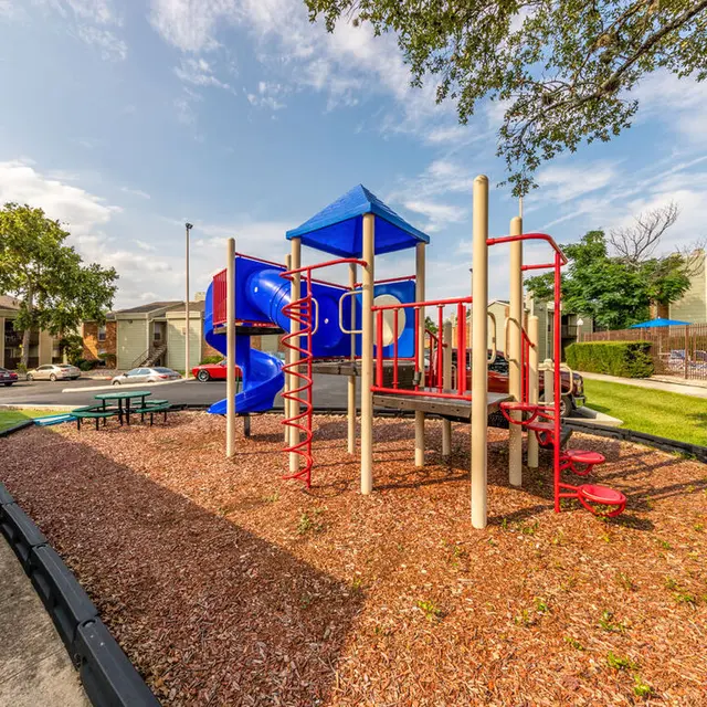 A colorful playground with a slide and climbing structures, surrounded by wood chips and green grass, under a blue sky with clouds.