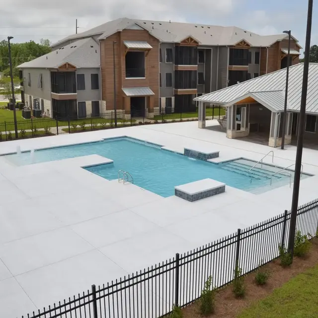 A view of a modern apartment complex with a swimming pool area and lounge space. The swimming pool is in the foreground, surrounded by a white concrete deck and fenced by a black railing. There are outdoor lounge chairs and a shaded pavilion nearby. The apartment buildings are three stories high with large windows and a contemporary design in the background.