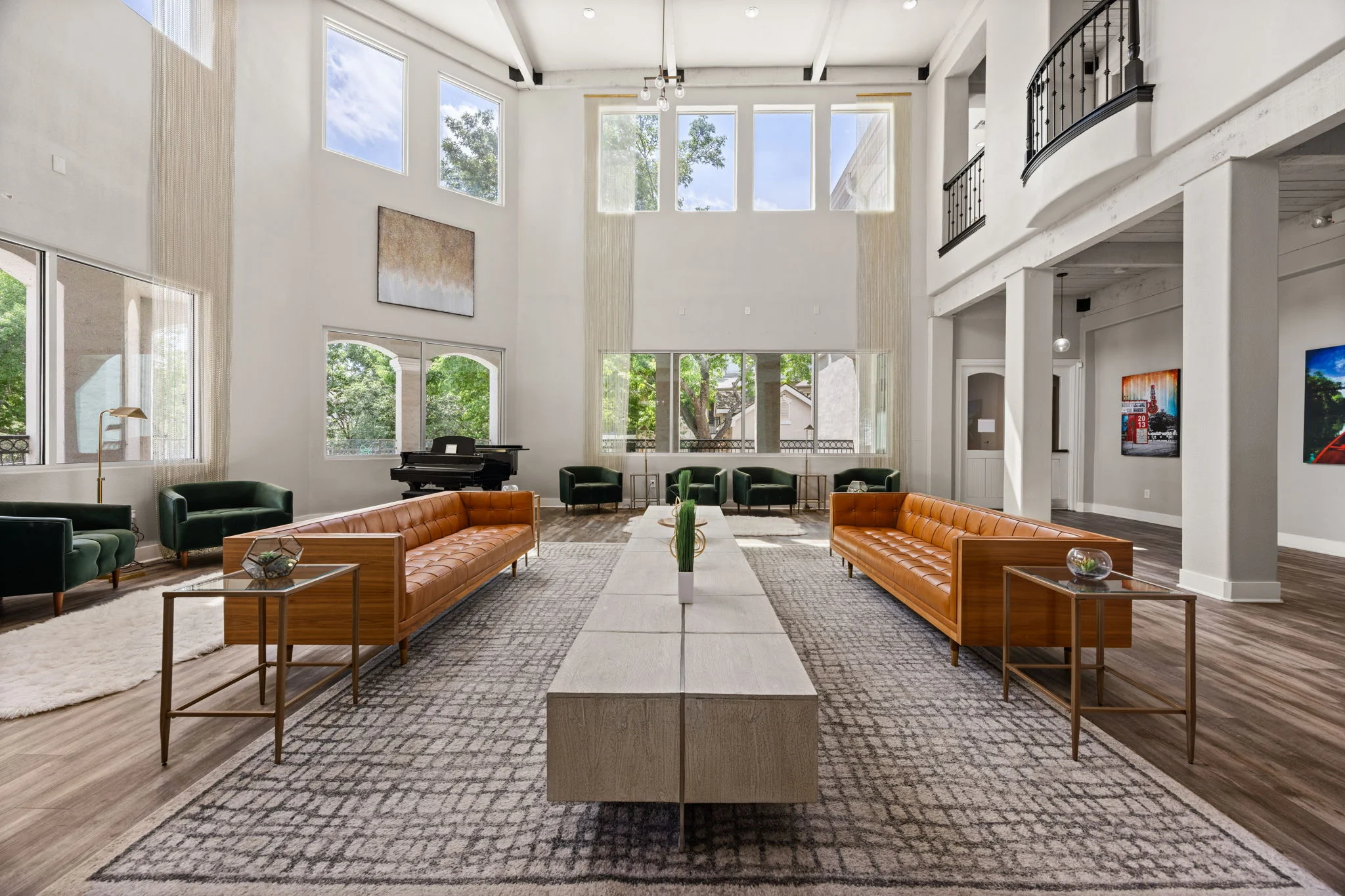 A spacious modern living room featuring two leather sofas, a long coffee table, and large windows allowing natural light in. There are green accent chairs and artworks on the wall, complemented by a soft area rug.