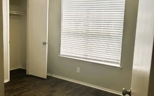 Empty room with light-colored walls, a window with blinds, a closet door open, and a wooden floor.