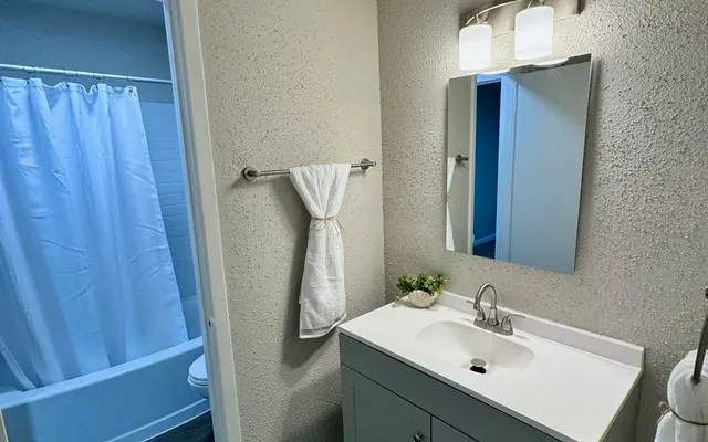 Modern Bathroom Interior A modern bathroom featuring a white cabinet sink, a large mirror, and a light fixture. A bathtub with a white curtain is visible in the background through an open doorway. The walls are painted in a light neutral color, and the floor has dark wood planks.