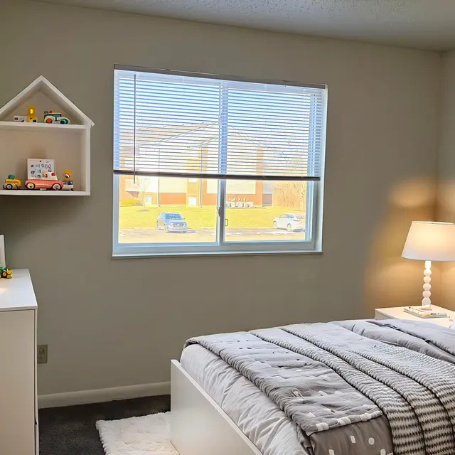 A cozy, well-lit bedroom featuring a bed with gray and white bedding, a white nightstand with a lamp, a shelf with decorative items, and a large window showing an outdoor view.