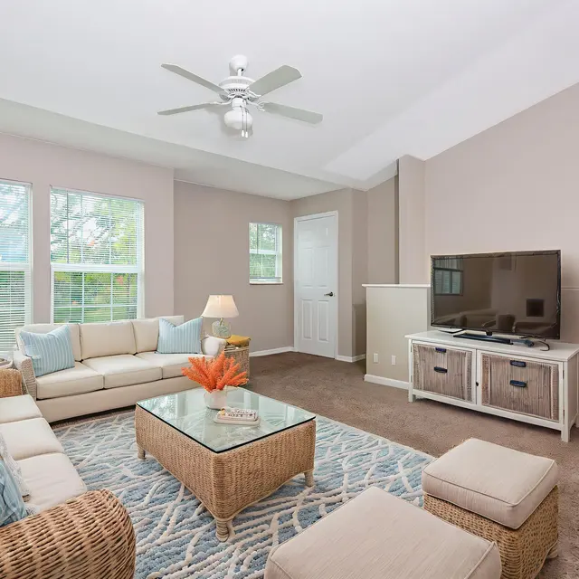 A cozy living room featuring a sectional couch, glass coffee table, and modern television. Large windows allow natural light, and a potted plant adds a touch of greenery.