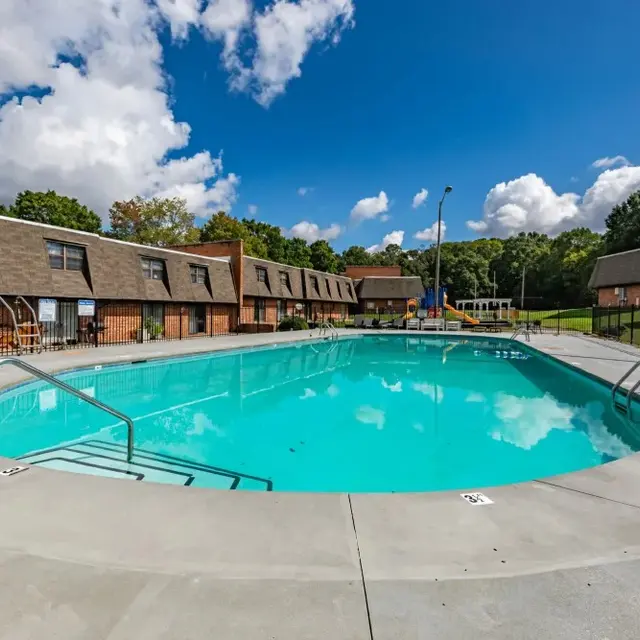 A clear blue swimming pool surrounded by a concrete deck and apartment buildings in the background, under a partly cloudy sky.