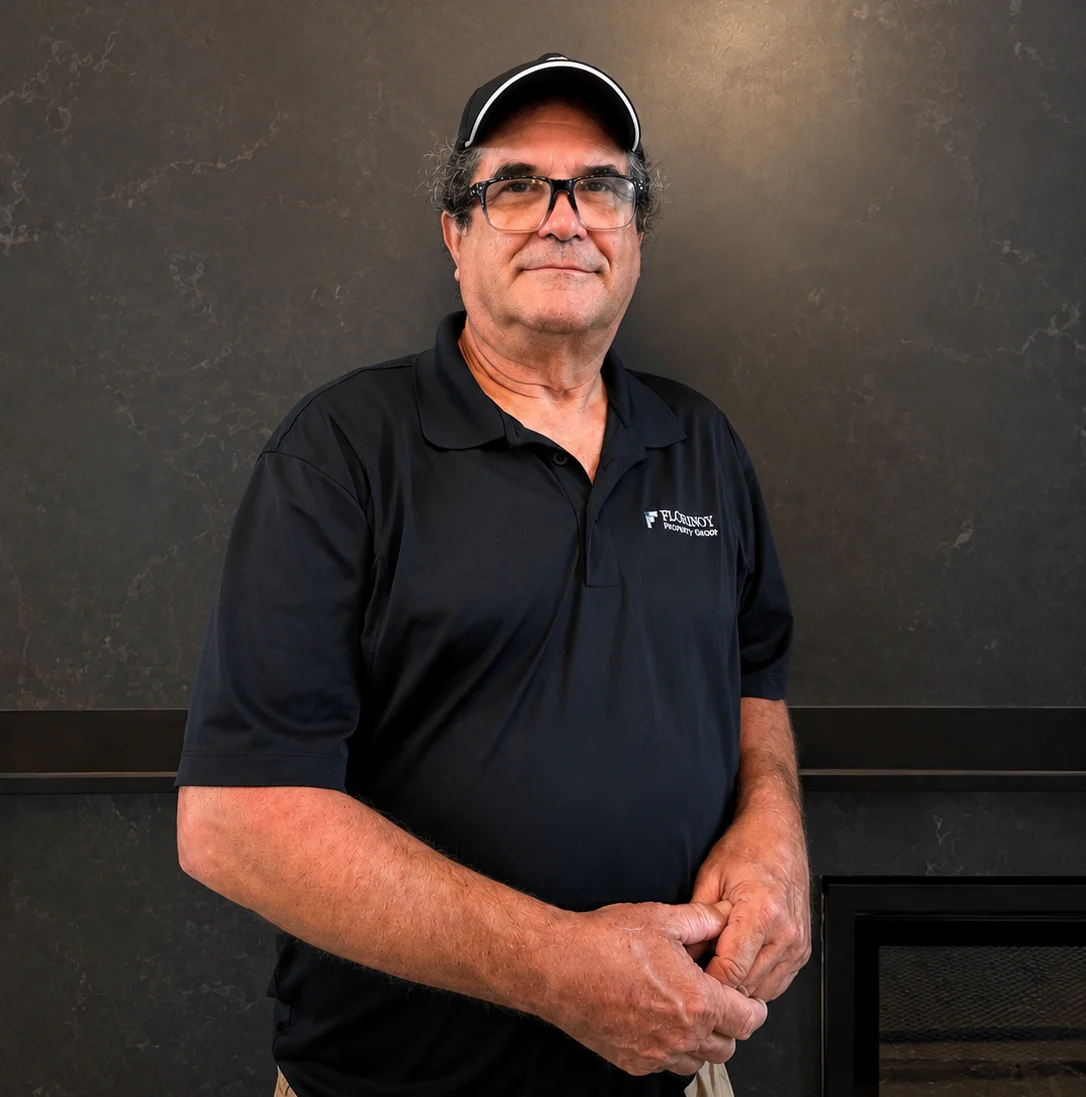 A man wearing a black polo shirt with a logo, standing against a textured dark wall.