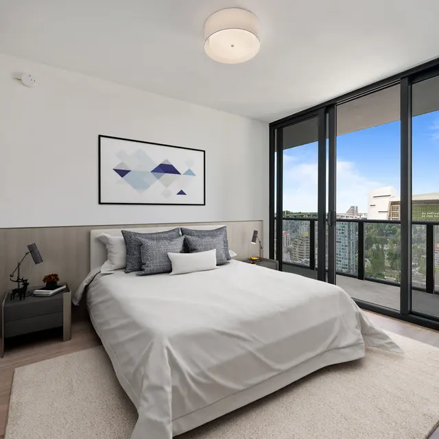 A modern bedroom featuring a queen-sized bed with neutral bedding, a decorative wall art piece, and large glass windows offering a city view.