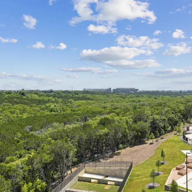 A panoramic view of a green landscape with trees, a blue sky with fluffy clouds, and a modern building on the right side.