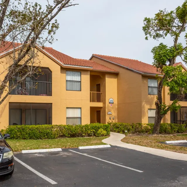 Exterior view of a two-story apartment complex featuring orange walls, a red tile roof, and surrounding greenery in the parking area.