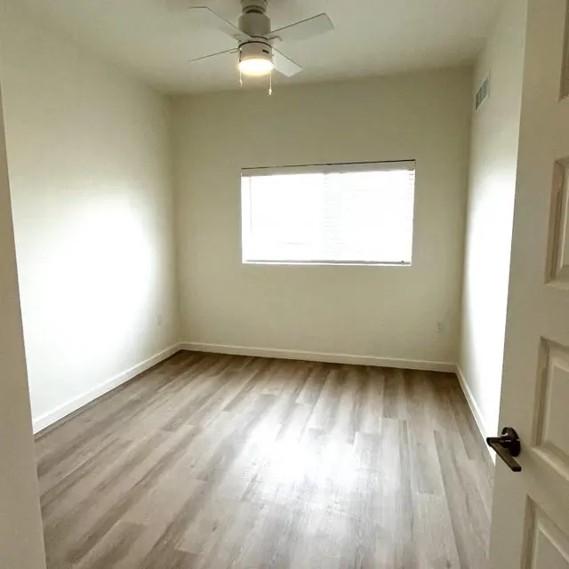 An empty room with light-colored walls and wooden flooring, featuring a ceiling fan and a window with blinds.