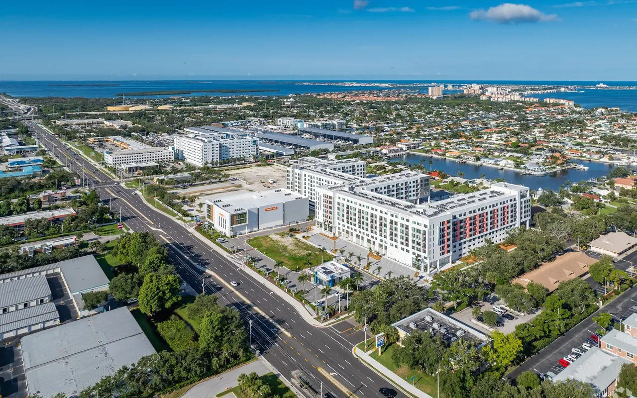 Aerial view of a modern urban area featuring residential and commercial buildings, green spaces, and water bodies in the background with clear blue skies.