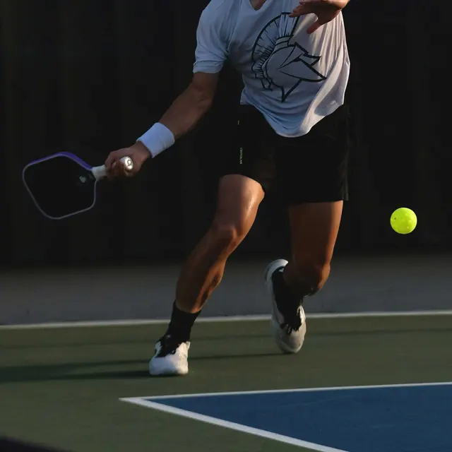 A player dressed in a white t-shirt and black shorts is in mid-swing, preparing to strike a yellow ball on a tennis court. The player is focused and in motion, with a paddle in hand and a backdrop of a tennis net and court surface.