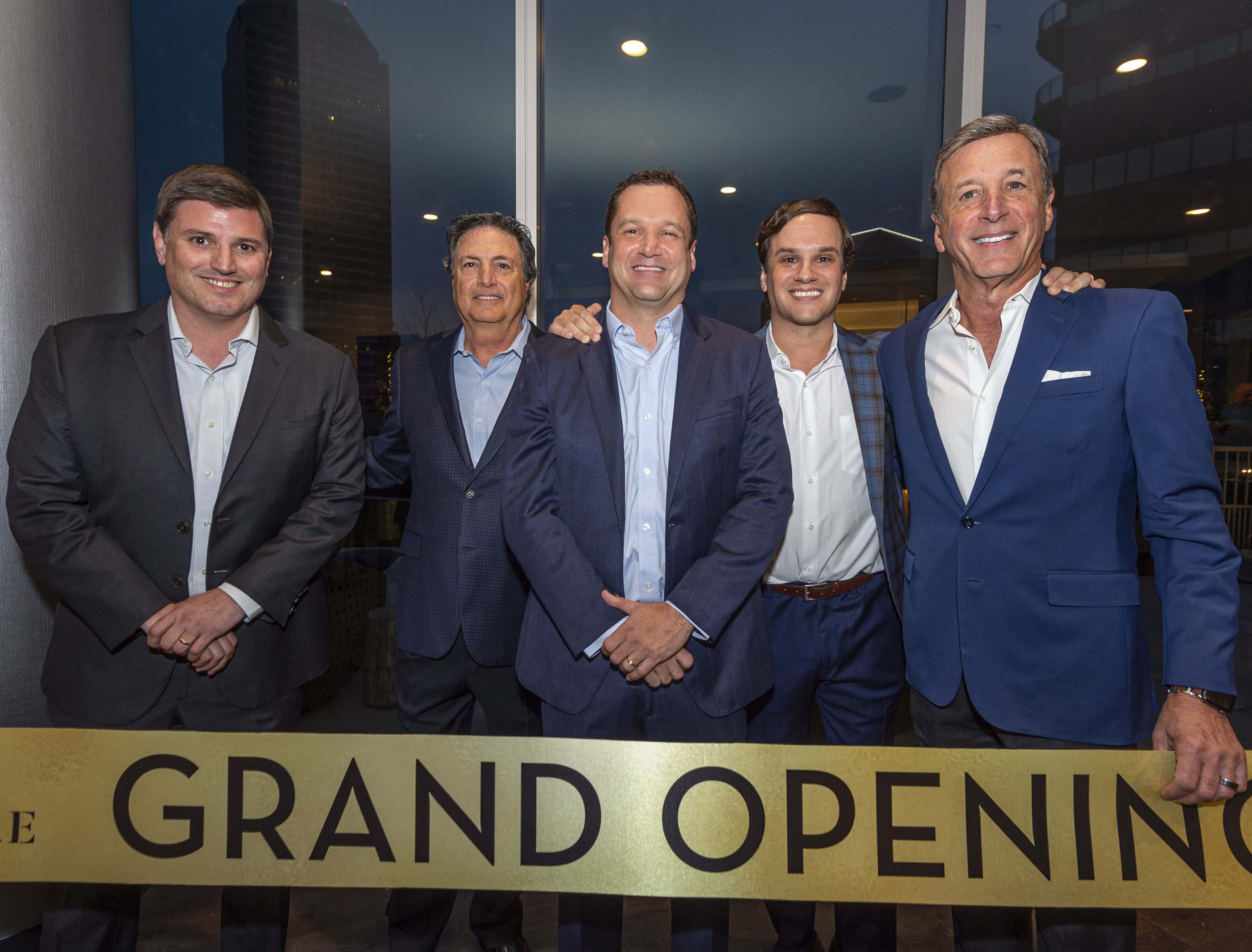A group of five men posing together at a grand opening event, standing in front of a large sign that reads 'GRAND OPENING.' They are dressed in business attire and smiling, with a cityscape in the background.
