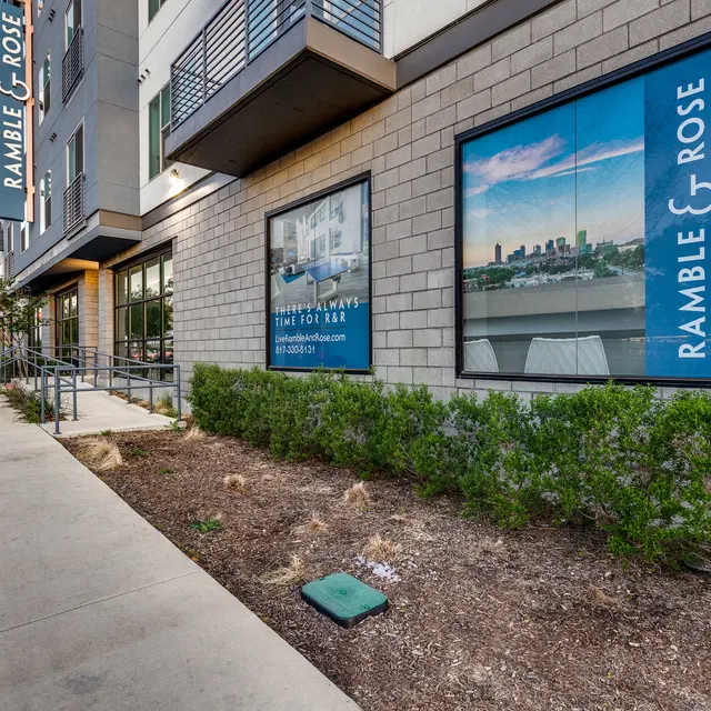 Exterior view of the Ramble & Rose building featuring promotional windows showing a city skyline and landscaped area with shrubs.