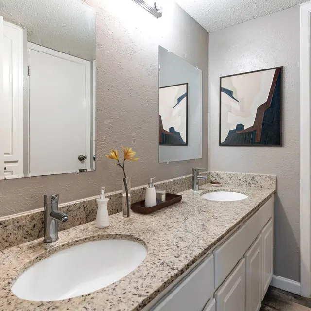 A bright, modern bathroom featuring two sinks with granite countertops, large mirrors above each sink, decorative flowers, and an adjoining area with a bathtub.