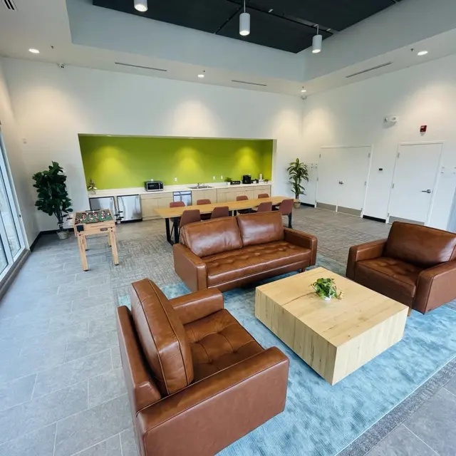 Modern Lounge Space A modern lounge area featuring brown leather sofas, a wooden coffee table, and green accent walls. Large windows allow natural light to fill the space, complemented by potted plants and a casual dining setup in the background.