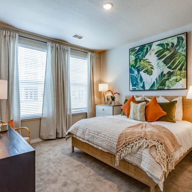Cozy Modern Bedroom Design A cozy modern bedroom featuring a warm color palette, with a large bed adorned with decorative pillows, a textured throw blanket, a piece of tropical leaf artwork on the wall, and natural light flowing in through large windows.