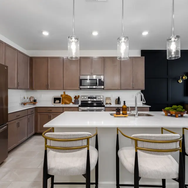 A modern kitchen featuring wooden cabinets, a stainless steel refrigerator, and a large white island with seating. Pendant lights hang above the island, and there is a stylish black wall with a TV mounted on it.