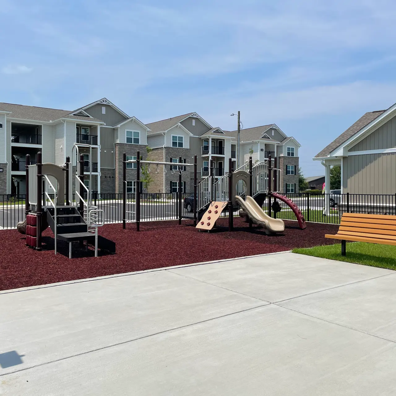 A playground area with slides and climbing structures, surrounded by a black fence and located next to residential buildings. There is a concrete space in the foreground and a bench on the side.