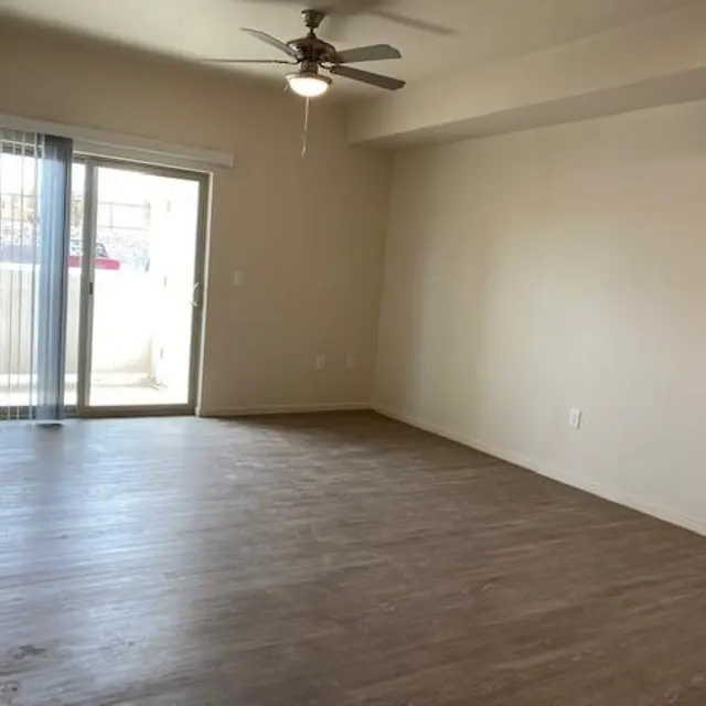 An empty living room with a ceiling fan, wooden floor, and a sliding glass door leading to a balcony.