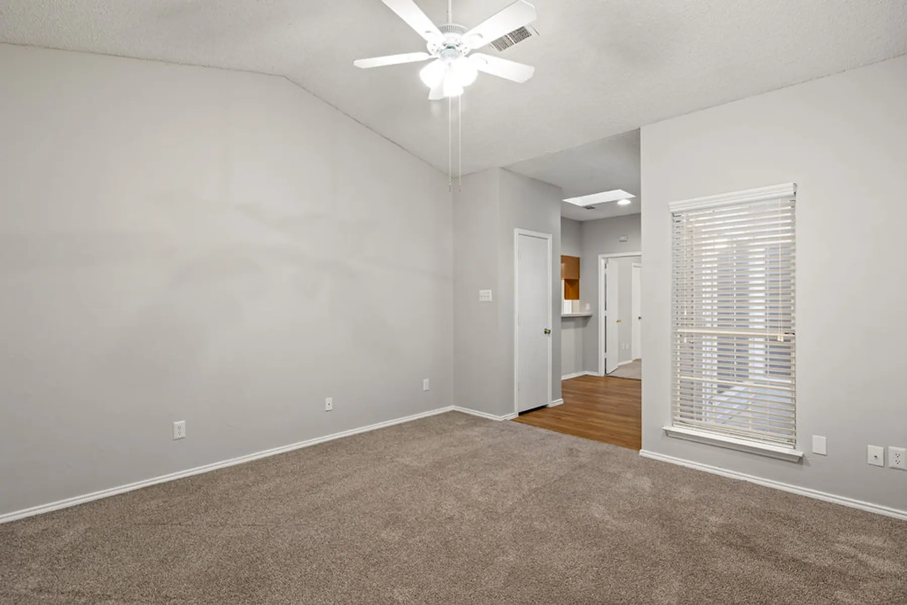 Spacious Living Room Interior A spacious living room with newly carpeted flooring, a ceiling fan, and light gray walls. There are two doors leading to other rooms, and a window with blinds letting in natural light.