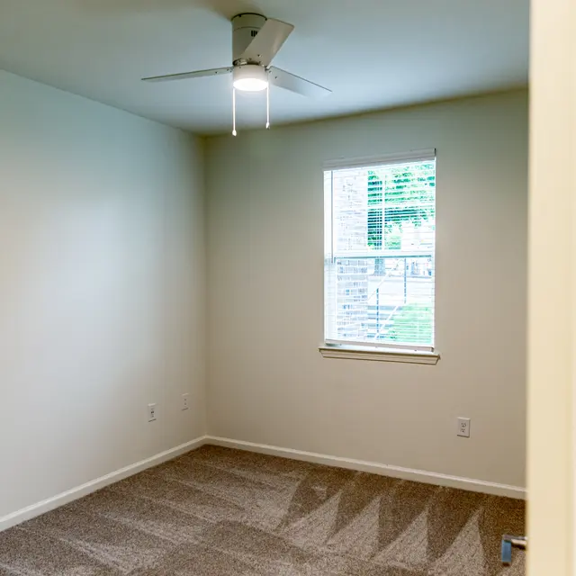 An empty room featuring a light-colored wall, window with blinds, and carpet flooring. A ceiling fan is mounted on the ceiling.