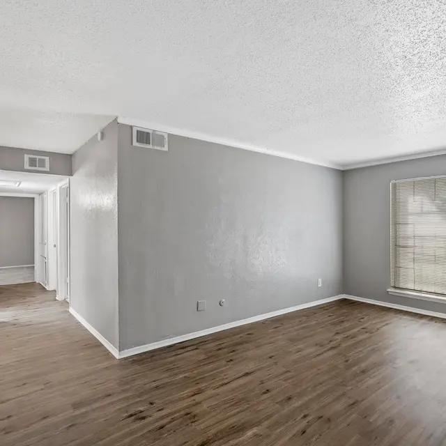 A spacious living room featuring light gray walls and a large window allowing natural light to enter. The floor has a wooden finish, and there are doorways leading to other rooms.