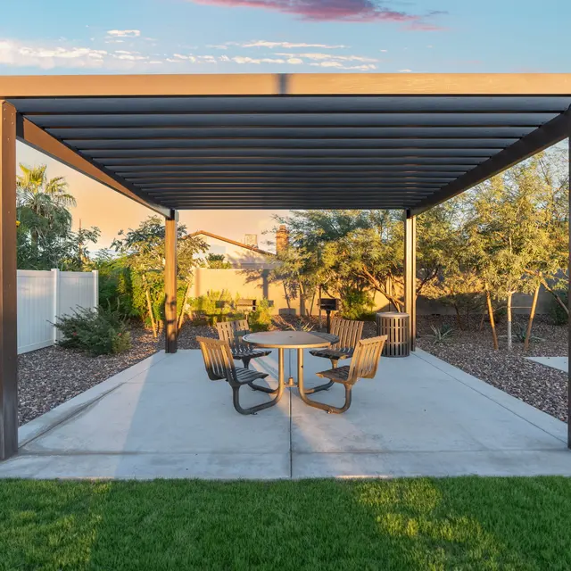 Modern Outdoor Pavilion A modern outdoor pavilion with a slatted roof, featuring a round table and several chairs on a concrete slab surrounded by desert landscaping.