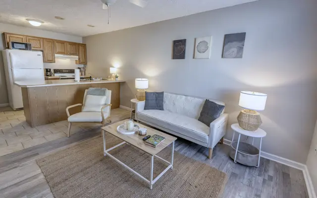 A modern living room featuring a white sofa with gray cushions, two lamps on side tables, a coffee table with books and a decorative item, a chair, and a kitchen area in the background.