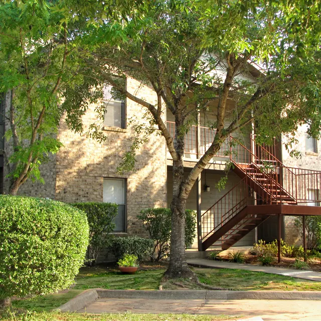A well-maintained brick apartment building surrounded by green bushes and trees. A staircase leads to the upper floor, and there are flower pots in the garden area.