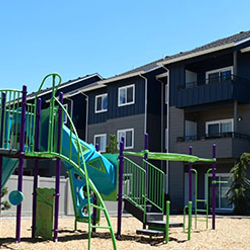 Playground and Apartment Complex A colorful playground featuring a blue slide and green play structures, set against a modern apartment building on a sunny day.
