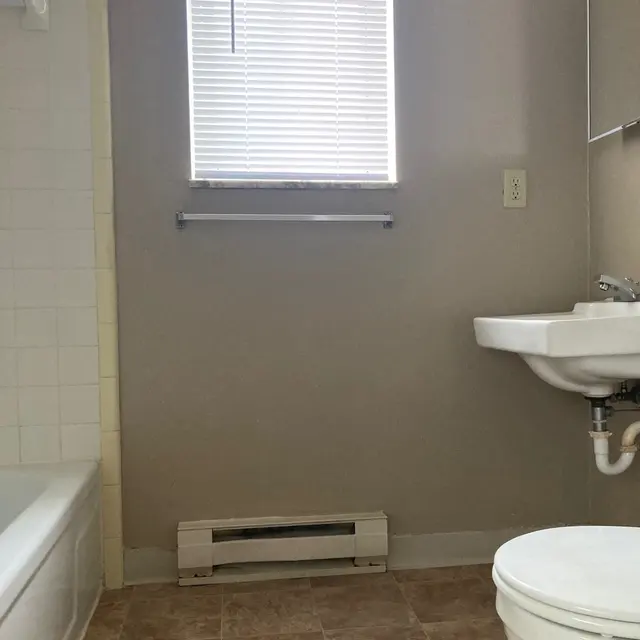 A clean bathroom featuring a bathtub on the left, a sink on the right, and a toilet in the foreground. The walls are painted a light grey, and there's a window with blinds letting in natural light.