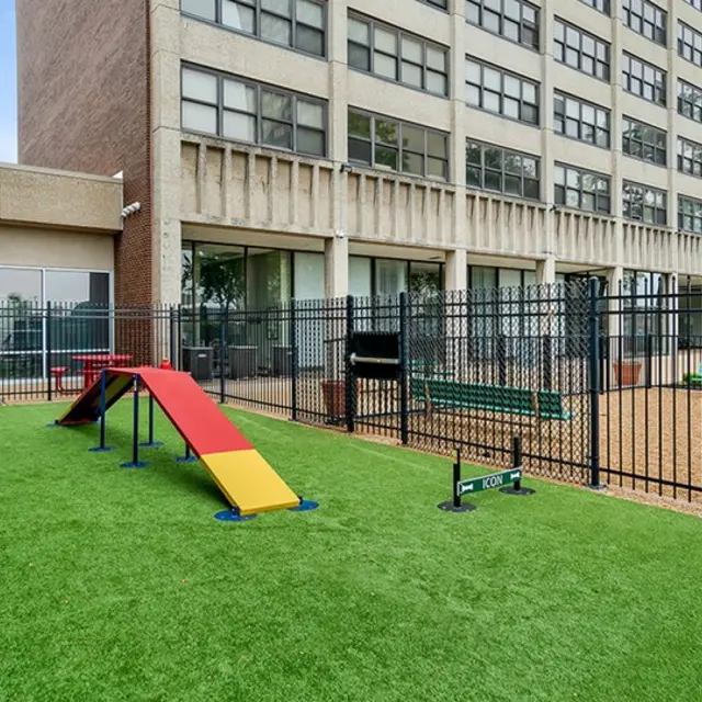 A fenced-in playground area featuring artificial grass, a red and yellow obstacle course, agility equipment, and playground structures. Behind is a multi-story building with many windows.