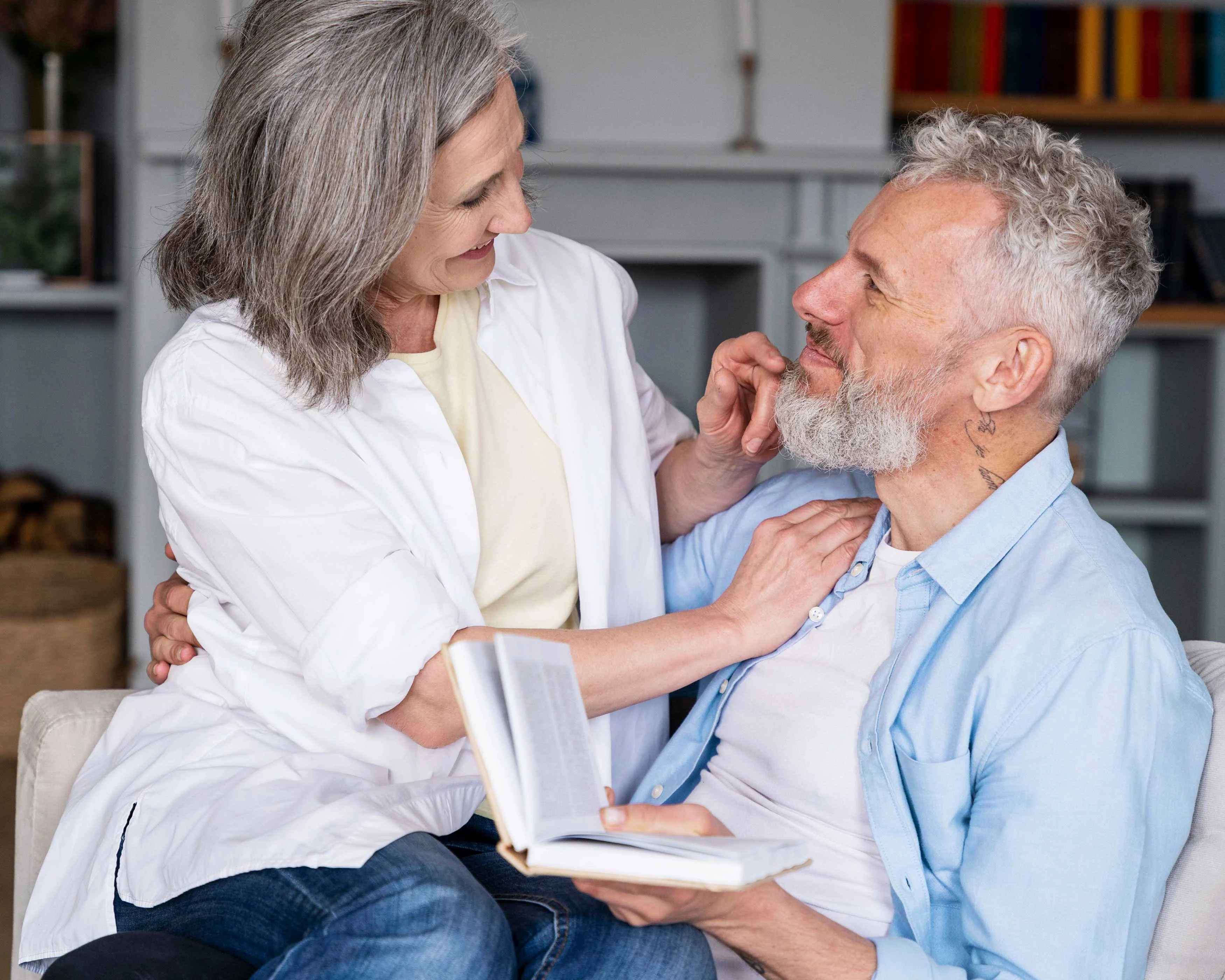 A joyful older couple cuddling on a couch, with a book in hand. The woman has gray hair and is wearing a white shirt, while the man has a beard and is wearing a light blue shirt. They are looking at each other affectionately.