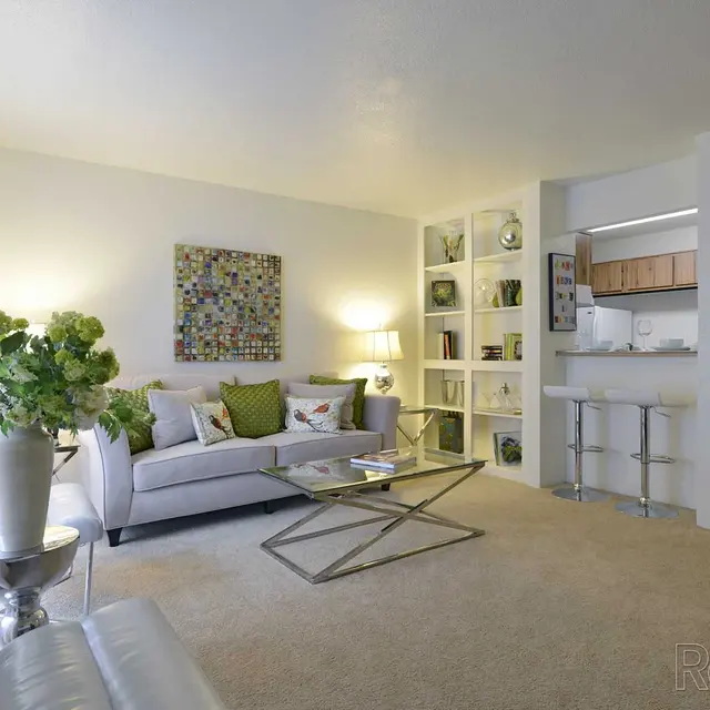 A contemporary living room featuring a light gray sofa with decorative pillows, a glass coffee table, and a small plant arrangement. A colorful art piece is on the wall, alongside a white bookshelf filled with decorative items. The room has large windows allowing natural light to enhance the cozy atmosphere.