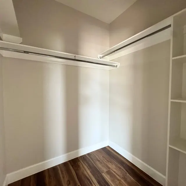 A spacious empty closet with a light gray wall, two hanging rods, hardwood floor, and a shelf on the right side.