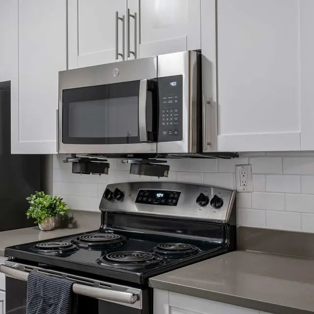 A modern kitchen featuring a black refrigerator, stainless steel microwave, and an oven with four burners. The cabinetry is white with silver handles, and there's a small plant on the countertop. The backsplash is made of white tiles.