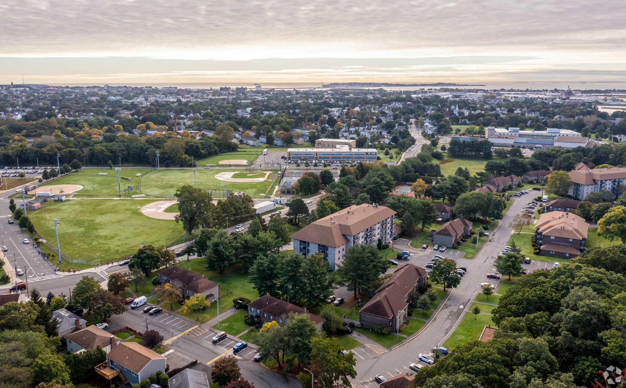 Aerial View of Suburban Neighborhood and Baseball Fields Aerial view of a suburban area featuring multiple baseball fields, residential buildings, and tree-lined streets. In the background, a city landscape is visible with a cloudy sky above.