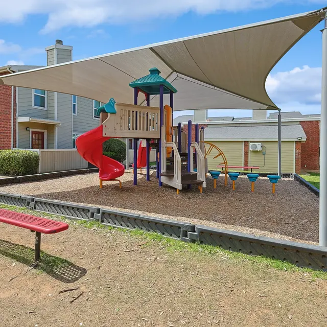A colorful playground with a slide and climbing structure, covered by a shade sail, located in a grassy area near residential buildings.