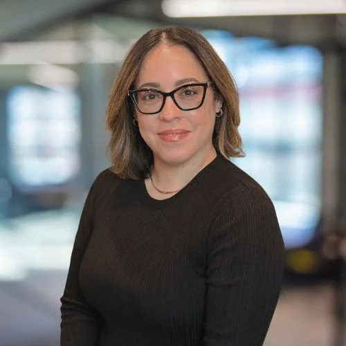 A woman with long curly hair wearing glasses and a black top, smiling and posing for a professional photograph. The background is blurred with soft lighting, giving a modern office ambiance.