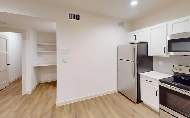 A modern kitchen featuring white cabinets, stainless steel appliances, and wooden flooring. A glimpse of a pantry area is visible on the left, with a doorway leading off to another room.
