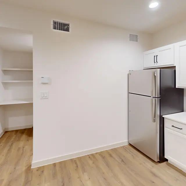 A modern kitchen featuring white cabinets, stainless steel appliances, and wooden flooring. A glimpse of a pantry area is visible on the left, with a doorway leading off to another room.