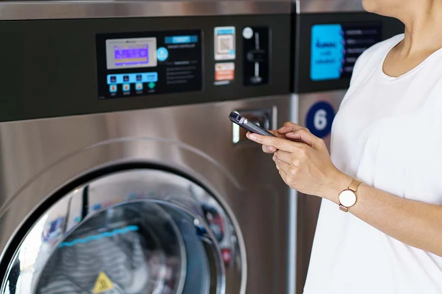 Tech-Savvy Laundry Experience A person standing next to a modern washing machine, holding a smartphone, with a digital control panel visible on the machine.