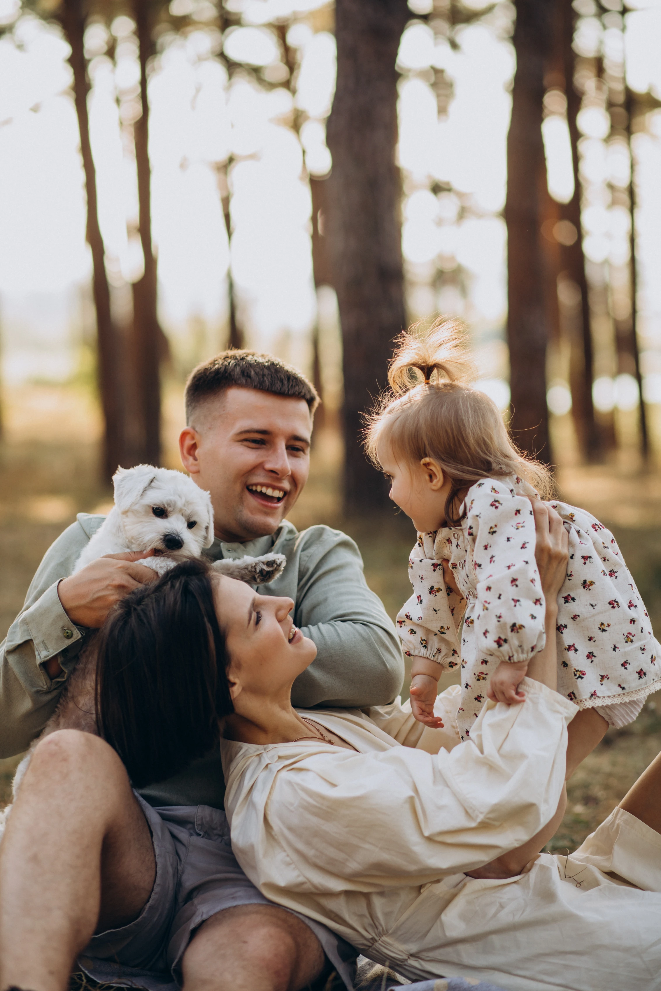 A joyful family outdoors with a child and a small dog. The father is holding the dog while the mother is interacting with the daughter, who is smiling and looking back at her mother. The setting is a sunny forest with tall trees in the background.