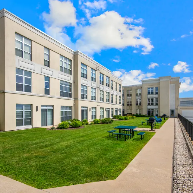 A modern multi-story building with large windows and green grass in front. The sky is bright blue with scattered clouds.
