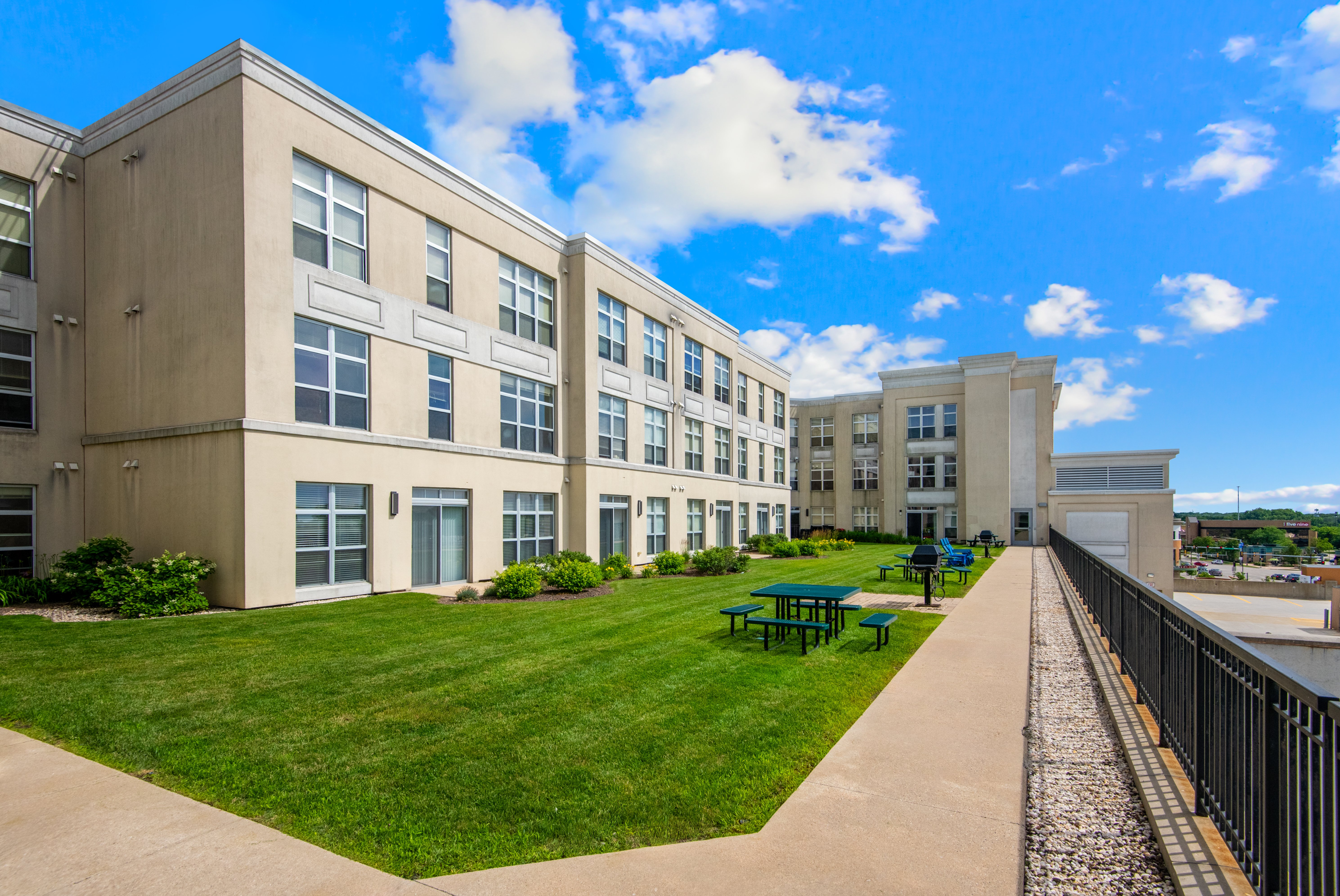 A modern multi-story building with large windows and green grass in front. The sky is bright blue with scattered clouds.