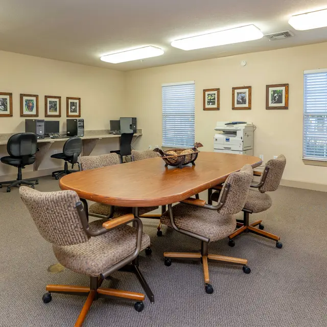 A spacious meeting room featuring a large wooden table surrounded by rolling chairs. There are computers on a counter along one wall, and a printer is positioned nearby. The room has large windows allowing natural light to enter, and framed artwork decorates the walls.