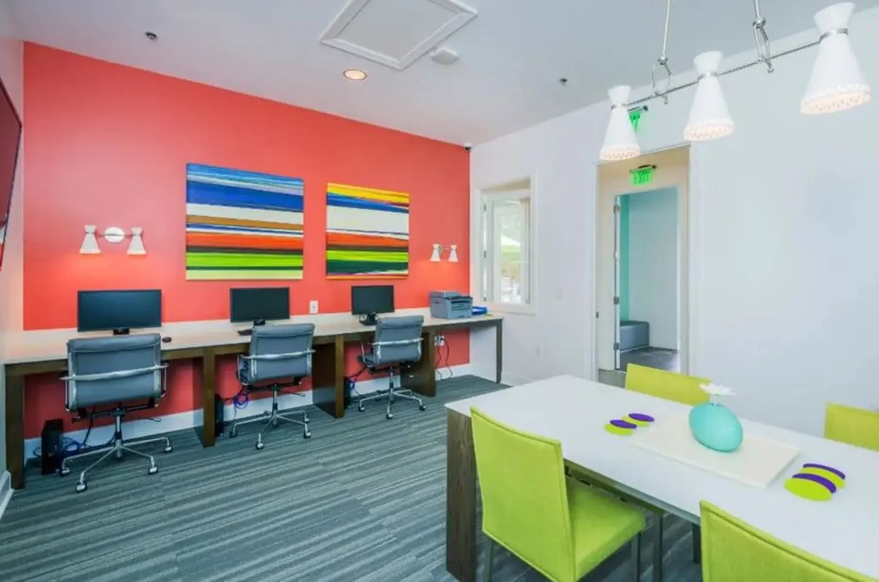 A modern office space with a vibrant red wall featuring abstract paintings. There are two workstations with computers and ergonomic chairs. A bright dining table with green chairs is in the foreground.