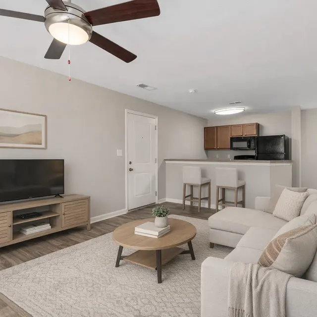 A modern living room featuring a light-colored sofa, a round coffee table with a plant on it, a television on a wooden stand, and a kitchen area in the background.