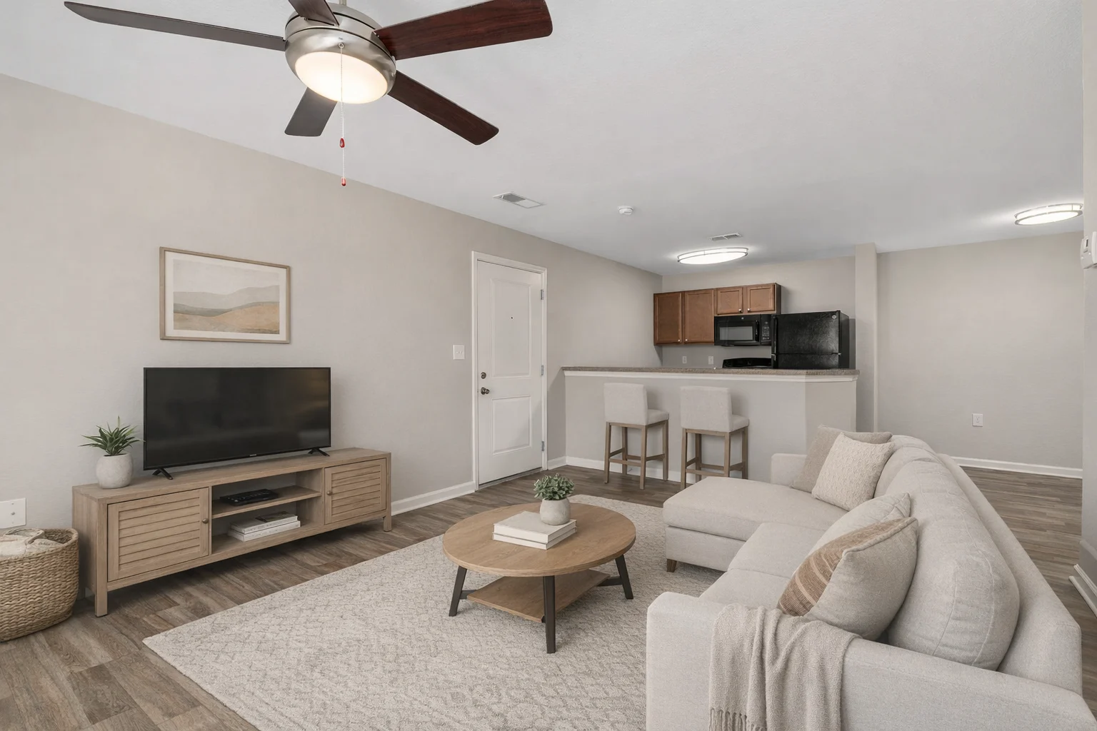 Modern Living Room with Open Kitchen A modern living room featuring a light-colored sofa, a round coffee table with a plant on it, a television on a wooden stand, and a kitchen area in the background.